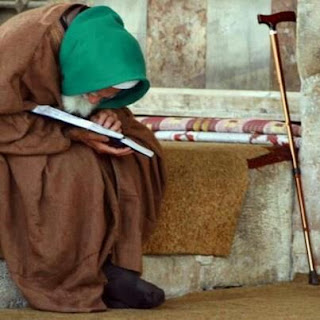 Seorang lelaki tua berserban hijau duduk membaca kitab di dalam masjid dengan tongkat di sisinya